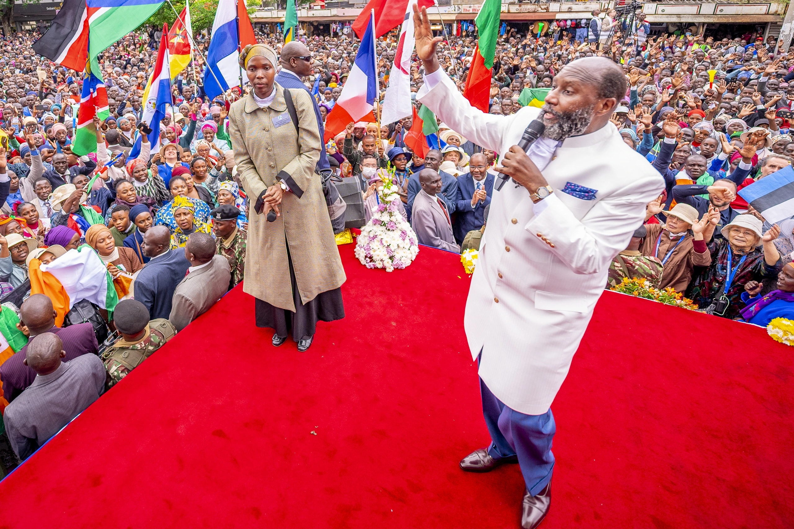 The preacher, in a white suit, raising his arm on a red platform in Nakuru, the flags of many nations and a vast crowd behind him.